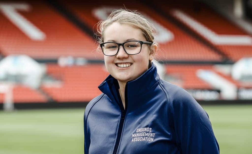 A person wearing glasses and a blue jacket smiles at a sports stadium.