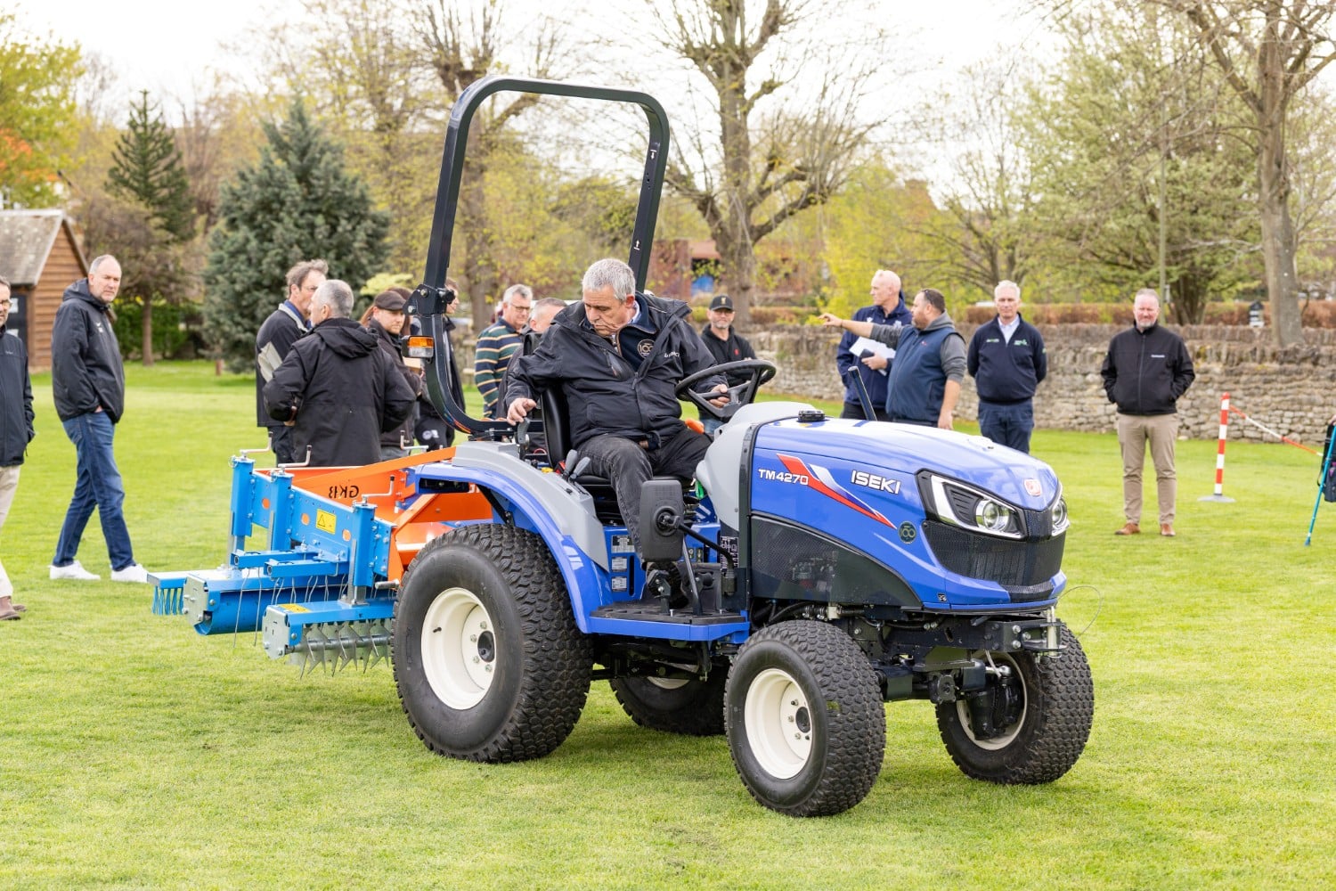 Man driving a blue tractor on grass with a group of people standing in the background.