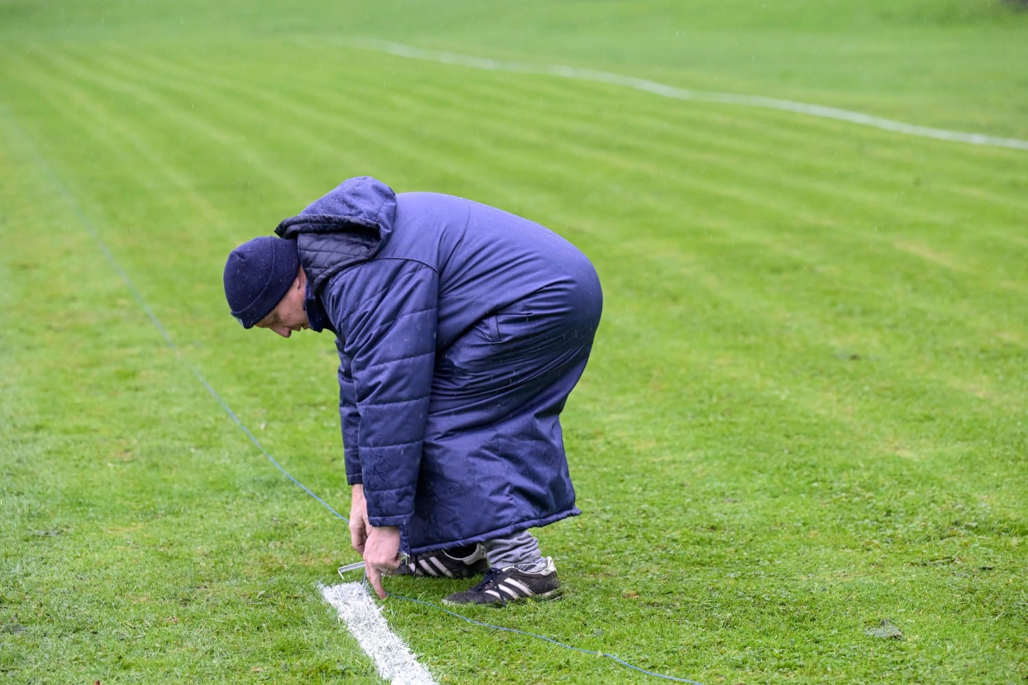 Person in a blue coat painting a white line on a grassy sports field.