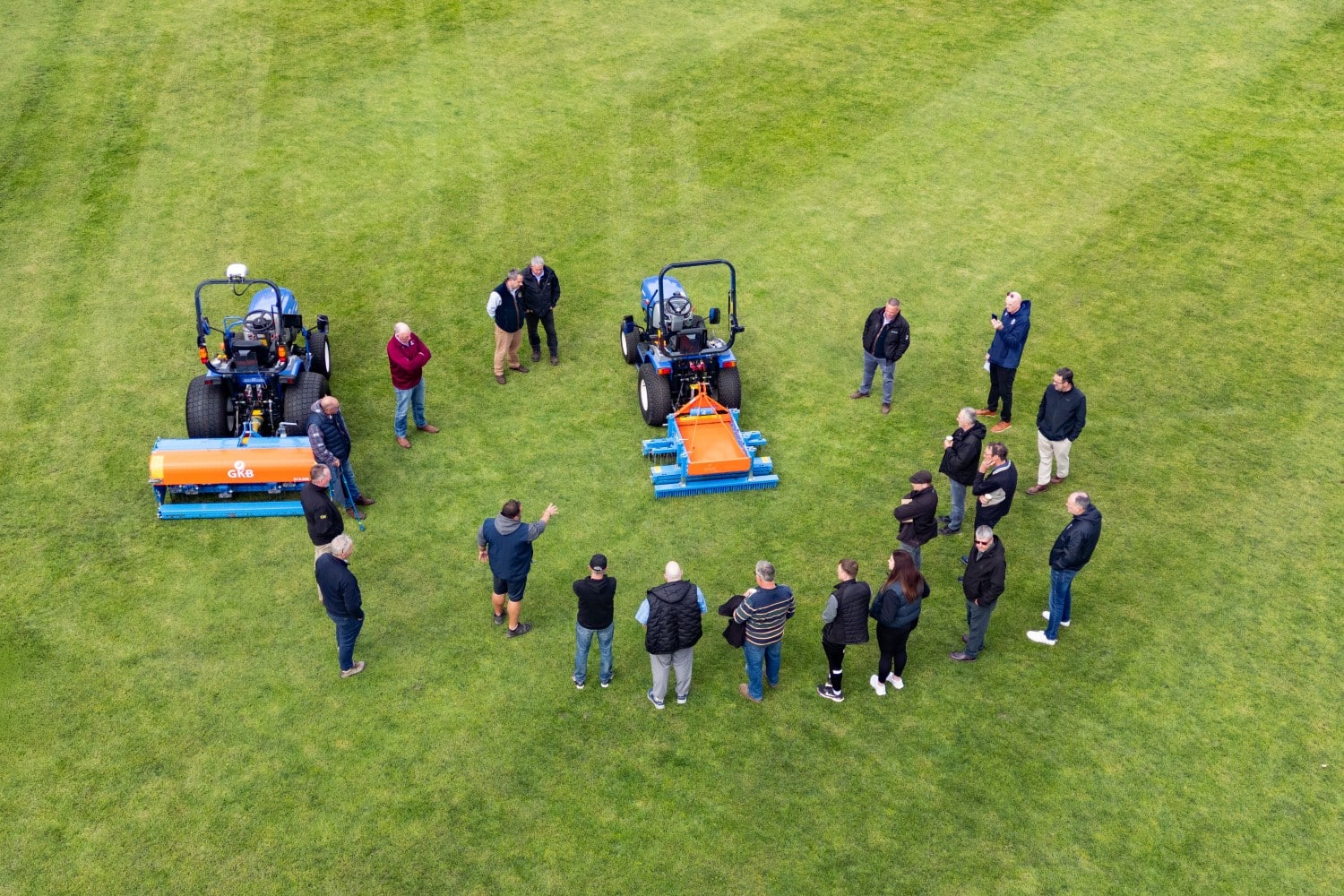 A group of people stand near two tractors on a grassy field, viewed from above.