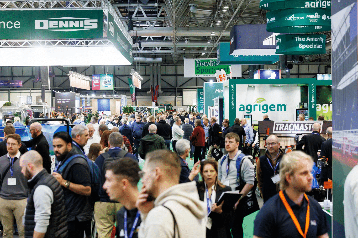 A large crowd of people attending an indoor trade show or exhibition with various company booths.