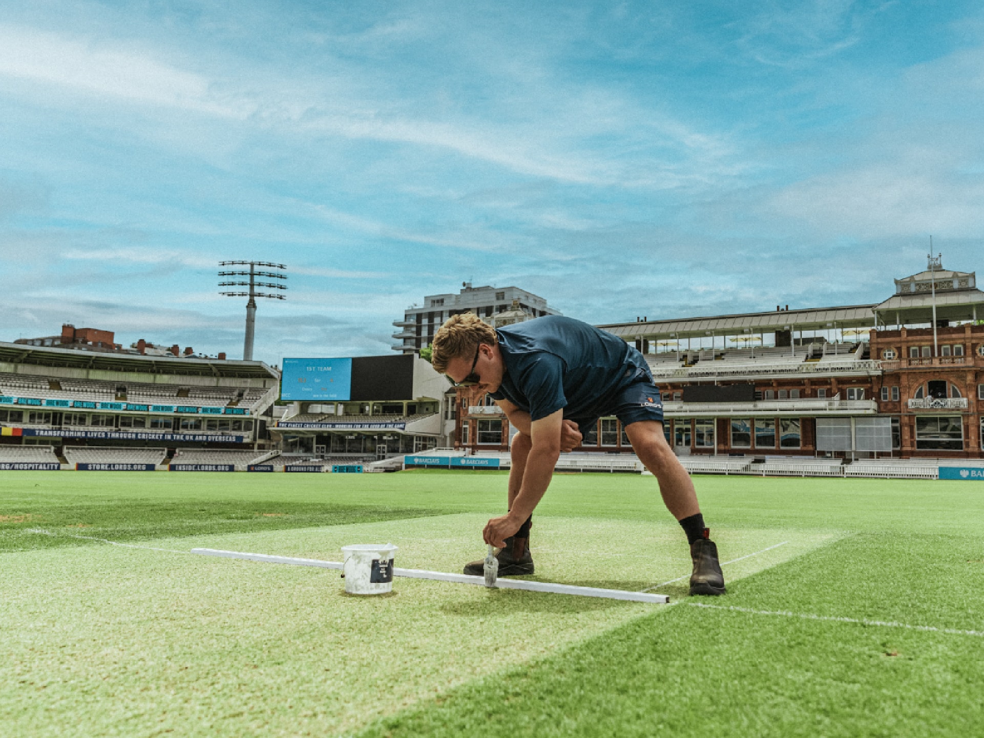 maintenance-funding-cricket Person painting a white line on a cricket field under a blue sky.
