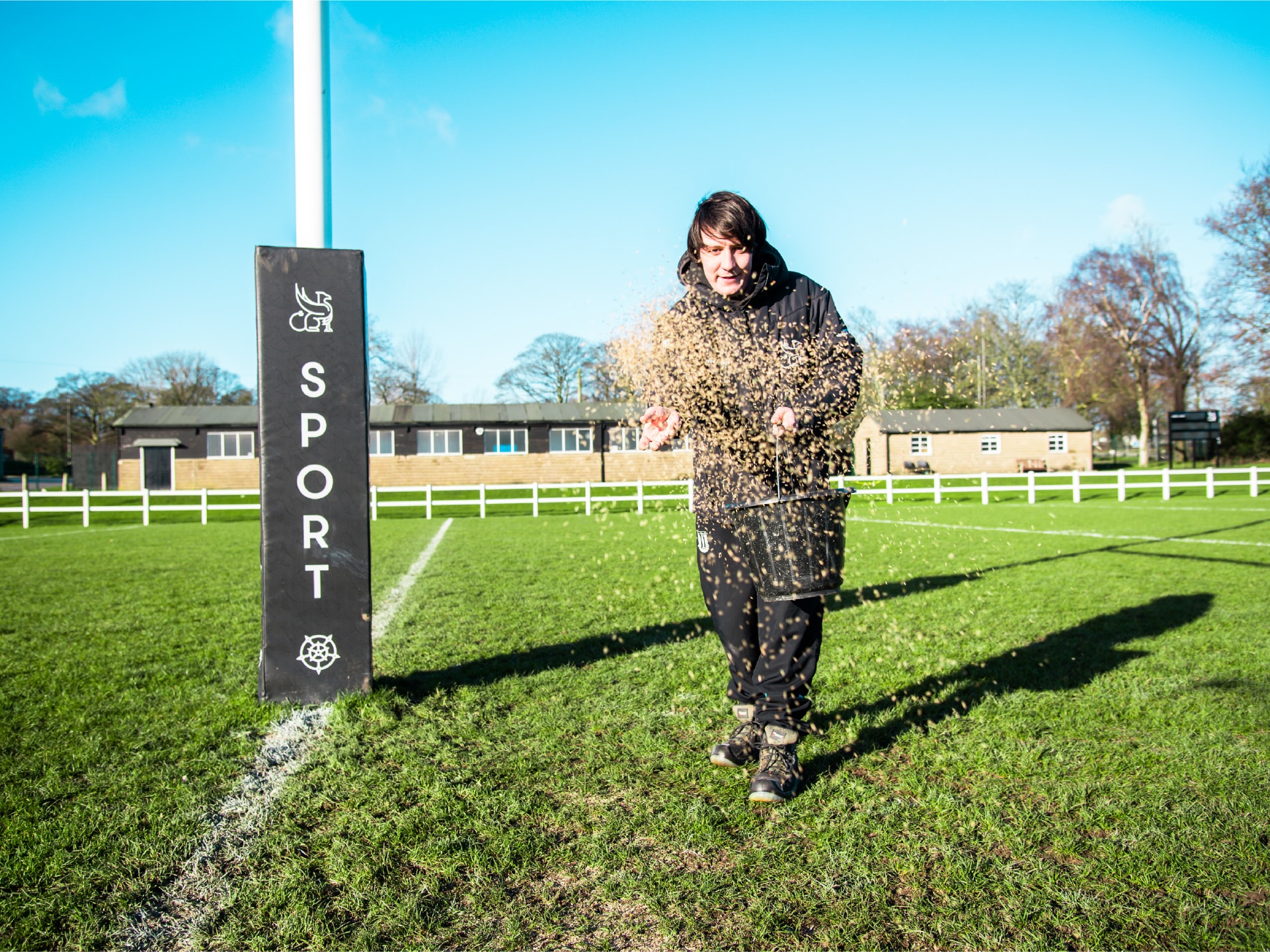 maintenance-funding-rugby-union Person spreading sand or soil on a rugby field near a goal post on a sunny day.